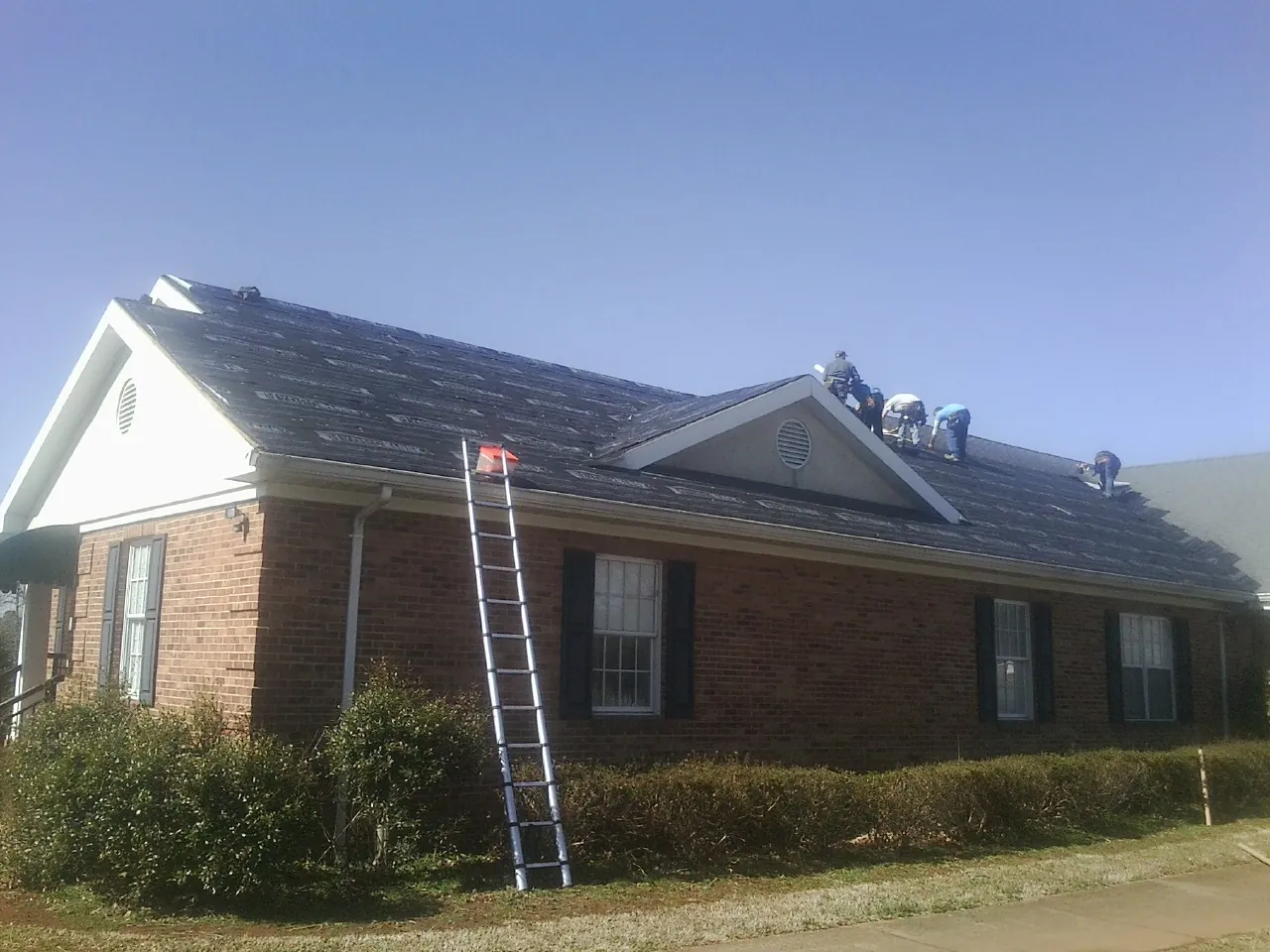Crew tearing shingles off a brick ranch roof with a ladder set — Emergency Roof Repair work by our crew in Lincroft