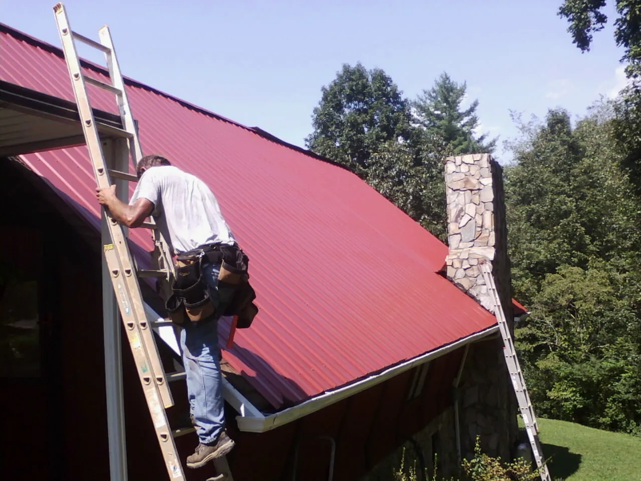 Roofer on a ladder beside a red metal roof and stone chimney — Soffit & Fascia Repair by Roofing Brothers in Lincroft